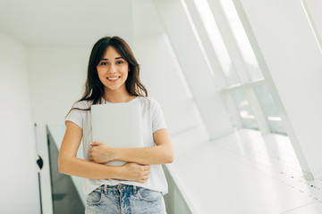 Fototapeta na wymiar Attractive cheerful young woman in casual clothes posing on camera and smile. Holding white laptop in hands. Stand in white room. Smiling alone.
