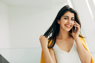 Fototapeta premium Picture of happy attractive young woman look straight on camera and smile. Touch hair and talk on phone. Businesswoman in deal. Stand at white wall or window.