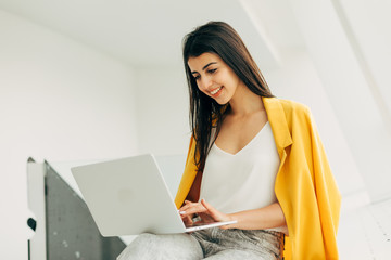 Busy cheerful young woman work in white room. Hold laptop on knees and typing on keyboard. Smiling...