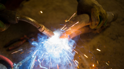 Fototapeta na wymiar A strong man is a welder in a black T-shirt, in a welding mask and welders leathers, a metal product is welded with a welding machine in the garage, blue sparks fly to the sides