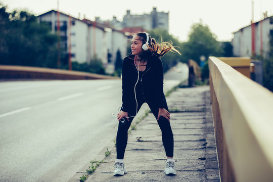 Young woman in black sports outfit resting while listening to music after running