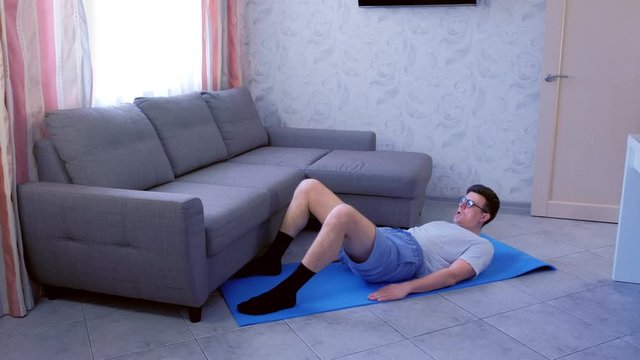 Funny nerd man in glasses is doing buttock bridge exercise on mat to strengthen the muscles of the legs at home. Sport humor concept.