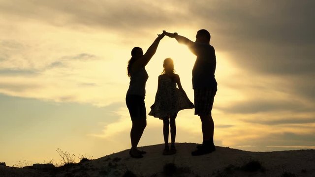 Beautiful family: mom, dad and little daughter on the background of sunset on the top of the hill. Parents build a house silhouette hands over the child's head. The concept of a happy family. 4K.