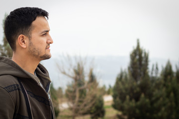 portrait of a young olive skinned man in sweater at the mountains surrounded by fir trees at midday in overcast day