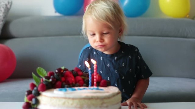 Beautiful two years old toddler boy in blue shirt, celebrating his birthday, blowing candles on homemade baked cake, indoors. Birthday party for kids
