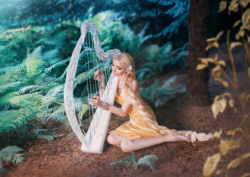 fabulous forest elf sits under tree and plays on white harp, girl with long blond hair braided in long yellow dress, summer goddess rests and sings to the sound of a magical musical instrument.