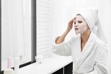 Beautiful woman with sheet mask on her face at mirror indoors