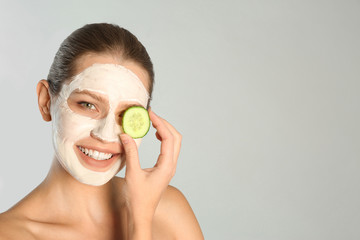 Beautiful woman holding cucumber slices near her face with clay mask against grey background. Space for text