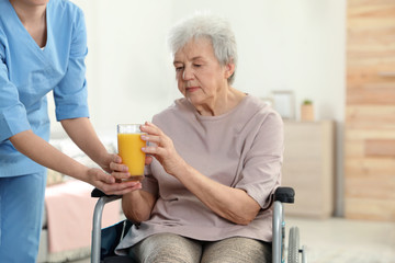 Nurse giving glass of juice to elderly woman in wheelchair indoors. Assisting senior people