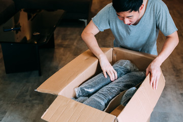 Young excited Asian man open box with product wrapped in plastic in living room