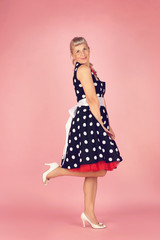Beautiful blonde woman in a polka dot dress, stands sideways and lifted her leg, pin-up style on a pink background