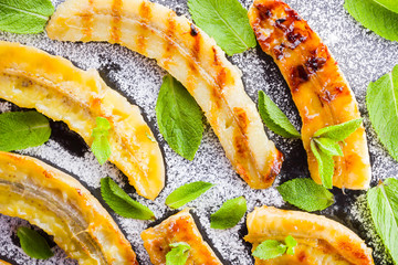 Halves of grilled bananas with powdered sugar on a black kitchen board and shale stone. Baked banana dessert with mint on black background table. Close up view © Lazartivan