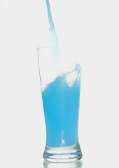 Liquido azul caindo em um copo de vidro, em a superficie branca e com fundo branco.