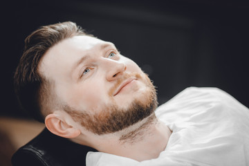 Fototapeta premium Man sits in barbershop chair and steam his face with hot towel in front of royal beard shaving razor