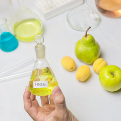 The lab assistent hand holds a flask of nitric acid on the background of the fruit lying on the...