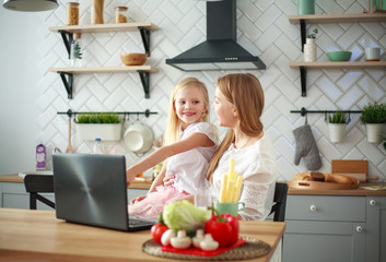 Mother with baby daughter sitting at table with groceries in kitchen with laptop