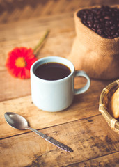 Black coffee In the cup sky blue color have roasted coffee beans are In a cloth bag sack butter cracker In the basket weave bamboo, still have red flowers and spoon all placed on a wooden table.