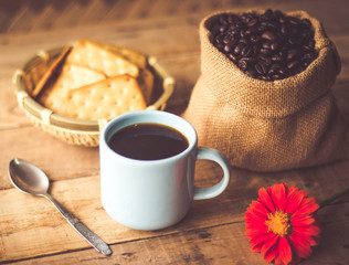 Black coffee In the cup sky blue color have roasted coffee beans are In a cloth bag sack butter cracker In the basket weave bamboo, still have red flowers and spoon all placed on a wooden table.