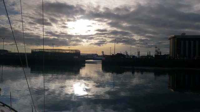 The sun behind clouds before setting with reflections in the water of a harbor