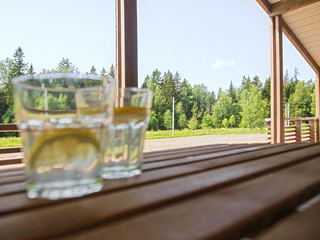 Wooden country furniture on the terrace of a country house. Two refreshing drinks out of focus with a refreshing lemonade with lemon slices in the foreground. Lush green foliage
