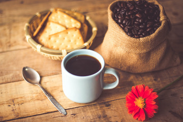 Black coffee In the cup sky blue color have roasted coffee beans are In a cloth bag sack butter cracker In the basket weave bamboo, still have red flowers and spoon all placed on a wooden table.