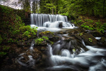 Amazing waterfall in the middle of mountain forest. River full of water during spring, beautiful flowing mass. Lovely contrast of water and surrounding forest green. Stones and fresh green leaves.