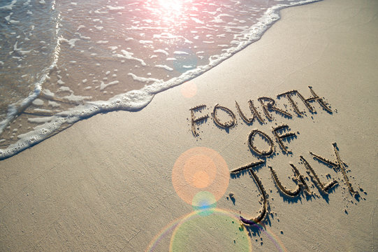 Fourth of July message written for the American Independence Day holiday in smooth sand with sunny lens flare view of an incoming wave on the beach