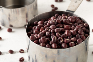 Raw, uncooked, dried adzuki (red mung) beans in metal measurement cups on white wooden table background