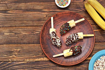 Dessert, frozen bananas in chocolate on a stick on a brown clay plate. Tropical desserts, party. Top view, copy space.