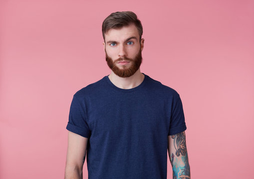 Photo of young handsome tattooed misunderstanding red bearded man in blank t-shirt, stands over pink background, looks at the camera with raised eyebrow.