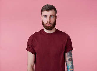 Portrait of sad young handsome red bearded tattooed man in red t-shirt, offended looking at the camera with a lowered lip, stands over pink background.