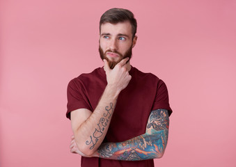 Portrait of young handsome thinking tattooed red bearded man in red t-shirt, looks away and touches...