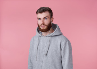 Portrait of young attractive red bearded man in gray hoodie, looks good and calm, smiles, istening song in headphones, enjoys the music, stands over pink background.