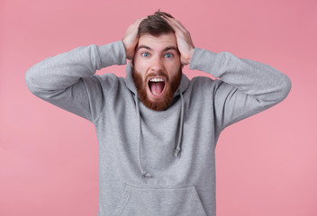 Portrait of young screaming shocked red bearded man in gray hoodie, looks evil and displeased , holds head, his favorite team lost. Stands over pink background and with wide open mouth.