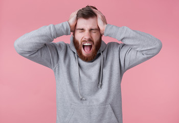 Portrait of young screaming shocked red bearded man in gray hoodie, looks evil and displeased , closed eyes andd holds head, stands over pink background and with wide open mouth