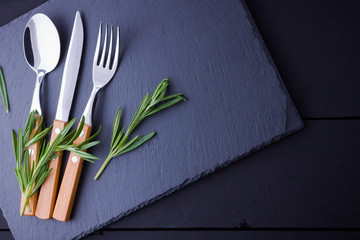 Set of cutlery knife, spoon, fork on slate stone. Сutlery with rosemary. Black background. Copy...