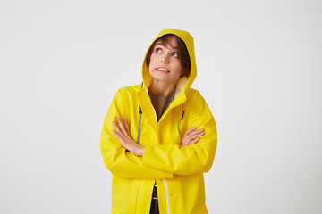 Portrait of young disgusted cute short haired girl wears in yellow rain coat, looks up with unhappy expressions, hiding under a rain hood, stands over white wall with crossed arms.