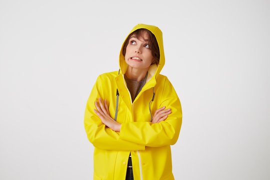 Portrait of young disgusted cute short haired girl wears in yellow rain coat, looks up with unhappy expressions, hiding under a rain hood, stands over white wall with crossed arms.