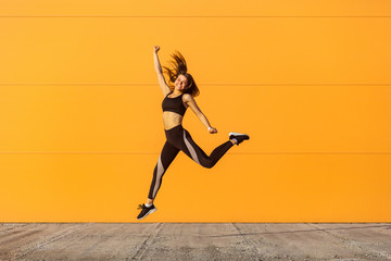 Young happy beautiful girl wearing black sporwear practicing sport exercises in morning on street, celebrate result of training, jumping full length, orange wall background, outdoor