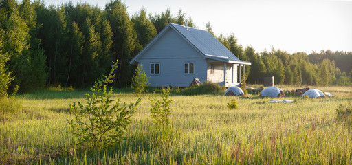 A painted one-story wooden house with a metal roof stands on the edge of the forest. There are scattered firewood nearby and there are small haystacks. Summer, dawn, Moscow region, Russia