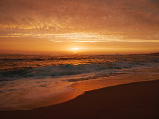 Waves crash on sandy beach at sunset with vivid orange sky