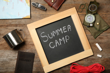 Chalkboard with text SUMMER CAMP and camping equipment on wooden background, flat lay