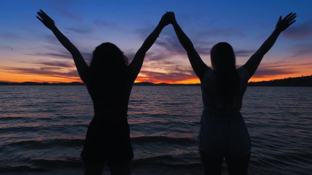Dolly, slow motion shot of a two girls holding hands, up in the air, standing on a beach, sunset in the background, at lake Huma, on a sunny evening dusk, at Hume Reservoir, New South Wales, Australia