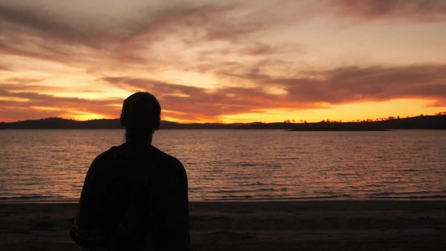 Handheld, slow motion shot of a woman standing on a beach, looking at the sunset, at lake Huma, on a sunny evening dusk, at Hume Reservoir, New South Wales, Australia