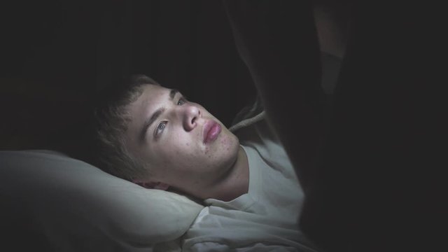 Male teenager lying on his bed while scrolling through social media on his phone. The camera pans into the darkness.