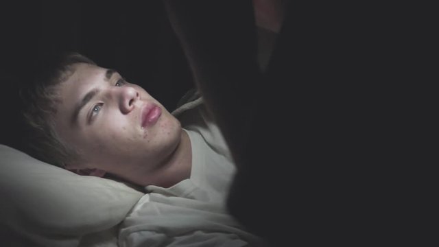 Male teenager lying on his bed while scrolling through social media on his phone. The camera pans in from the darkness.