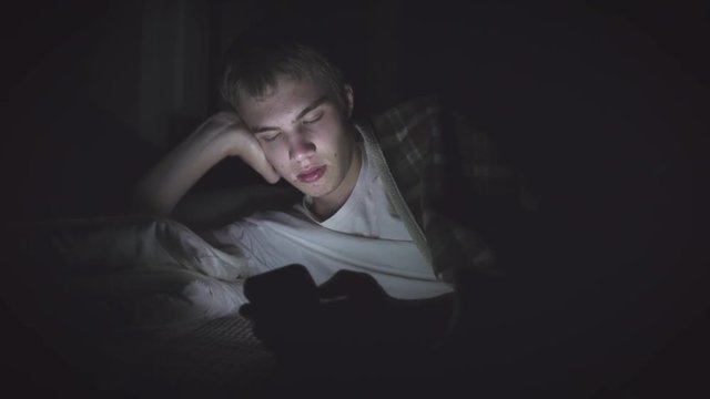 Bored teenager lying on his bed in the dark while scrolling through his phone. The light from the phone is illuminating his face.