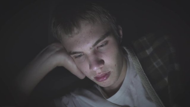 Bored teenager lying on his bed in the dark while scrolling through his phone. The light from the phone is illuminating his face.