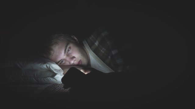 Bored teenager lying on his bed in the dark while scrolling through his phone. The light from the phone is illuminating his face.