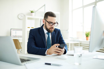 Serious concentrated young bearded businessman in formal suit sitting at desk in office and reading sms on smartphone 